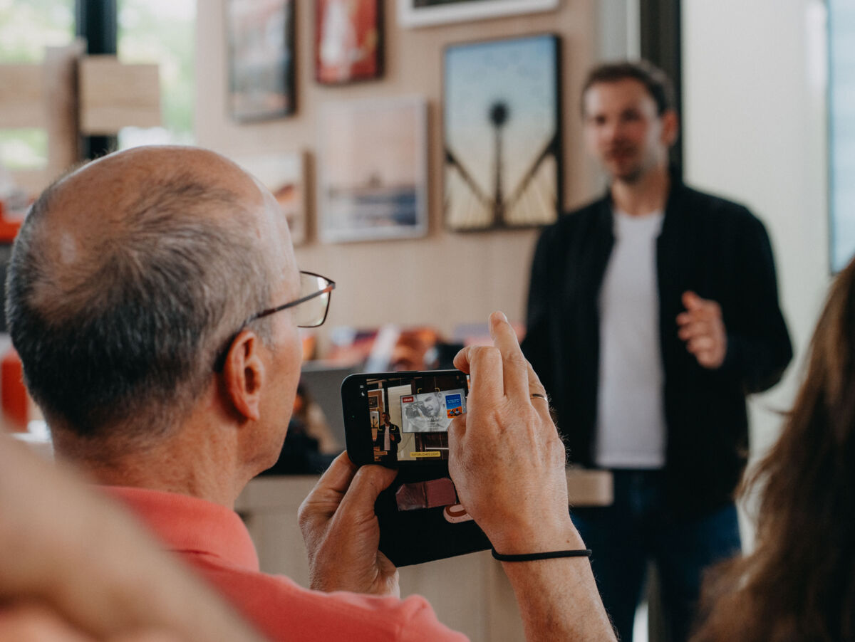 Ein Mann im Publikum fotografiert einen anderen Mann, der einen Vortrag hält.